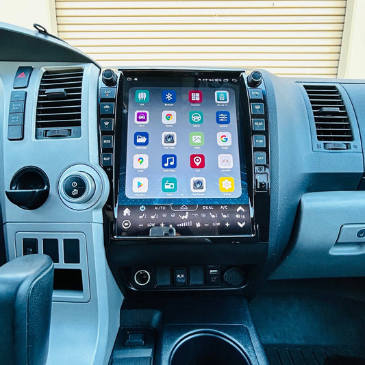A large touchscreen infotainment system mounted on the center console of a vehicle interior, showing a grid of app icons on the screen. The dashboard, steering wheel, gear shift lever, and center console are visible in gray and black tones.